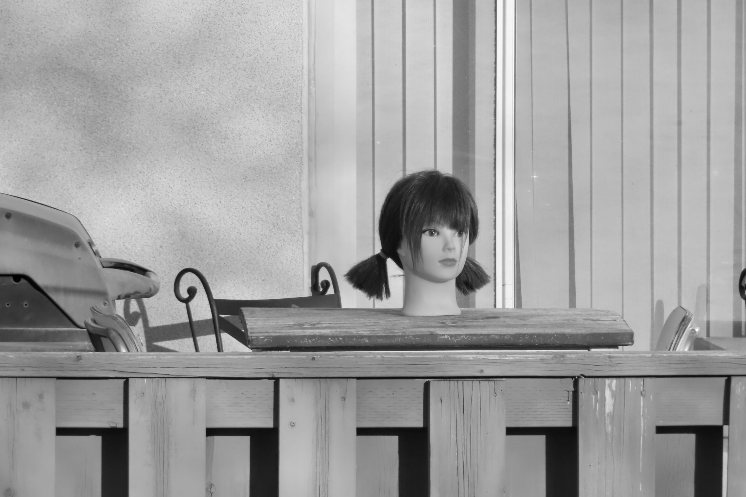 A mannequin head sits on an outdoor table behind a wooden fence. The photograph prompt for Sitting Still by Jonathan B. Edvane.